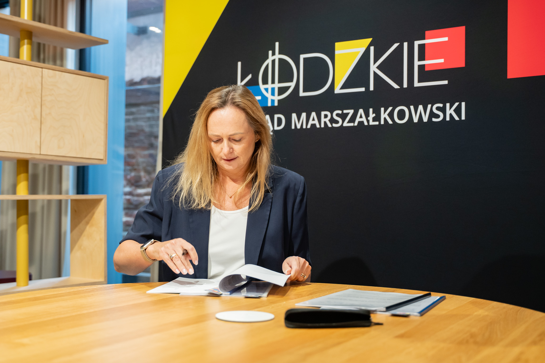 Joanna Skrzydlewska, marszałek Województwa Łódzkiego, przegląda dokumenty umowy przy stole na tle logotypu Łódzkie